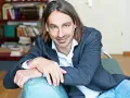 Richard David Precht sitzt lächelnd auf einem türkisfarbenen Sofa, im Hintergrund ein Bücherregal.