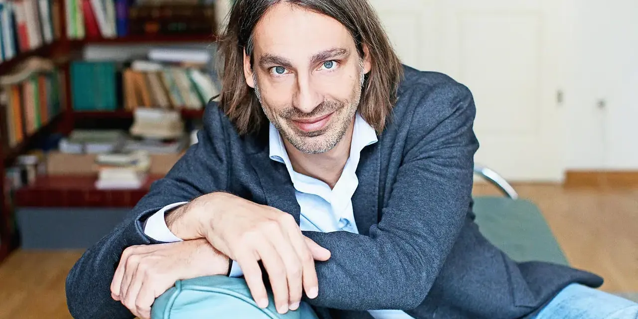 Richard David Precht sitzt lächelnd auf einem türkisfarbenen Sofa, im Hintergrund ein Bücherregal.