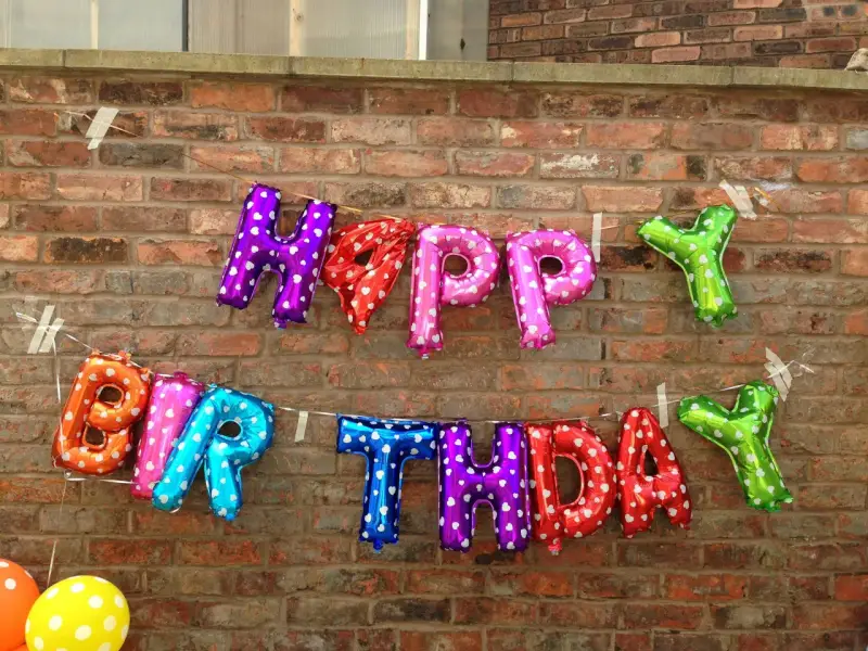 Luftballons in Buchstabenform bilden "HAPPY BIRTHDAY" an einer Ziegelwand.
